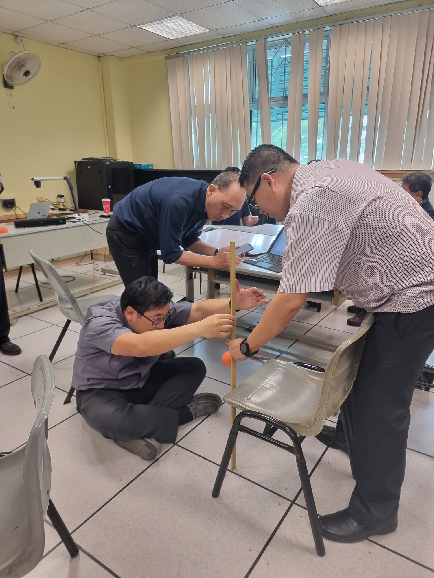 Three men in a classroom measure a vertical stick near a chair, one sitting on the floor holding an orange ball.